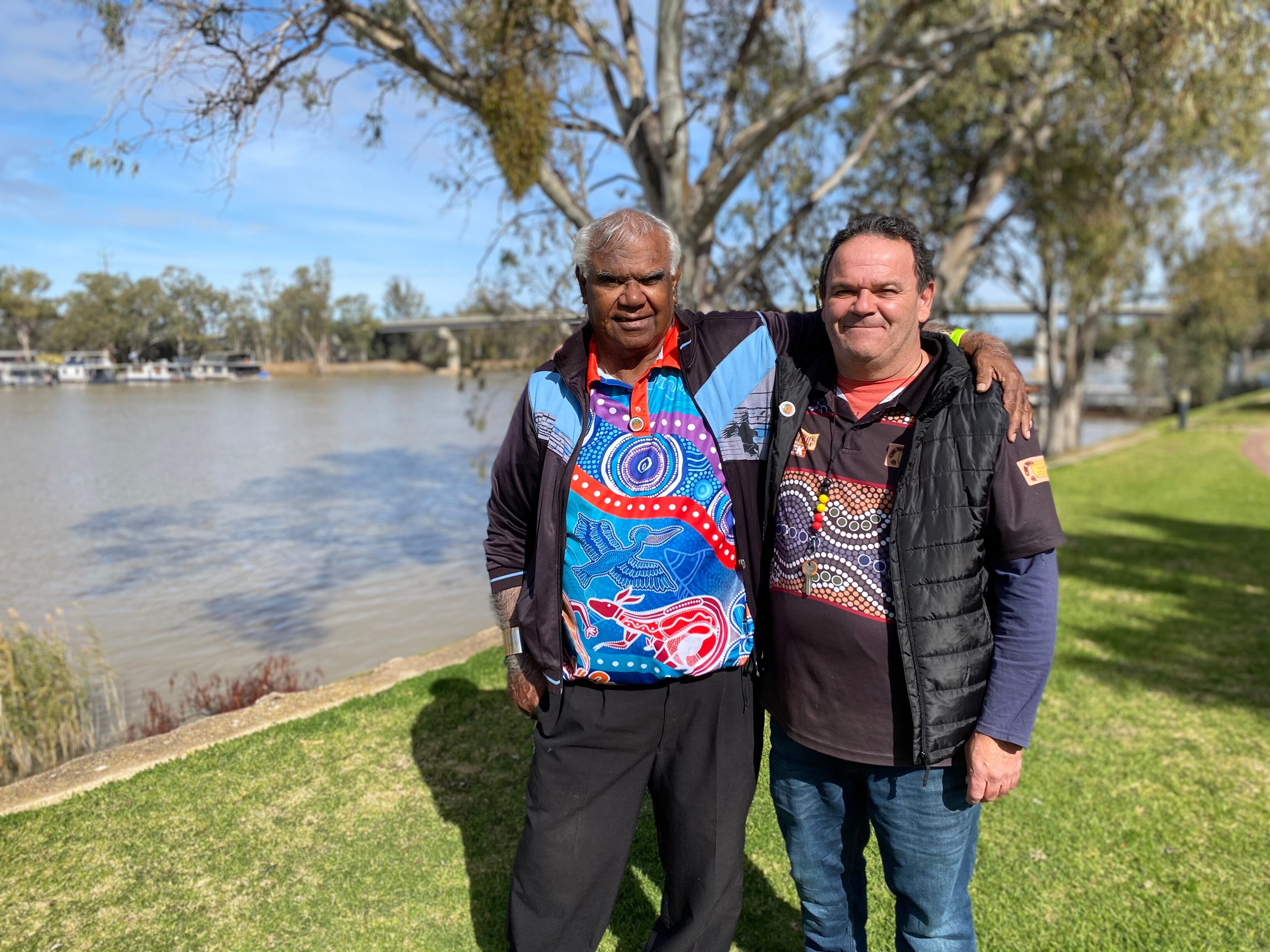 Two men stand together with arms wrapped around each other. They're standing in front of a river.