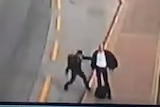 A screen shot from security vision shows a man swinging at another man standing at a bus stop as traffic passes by.