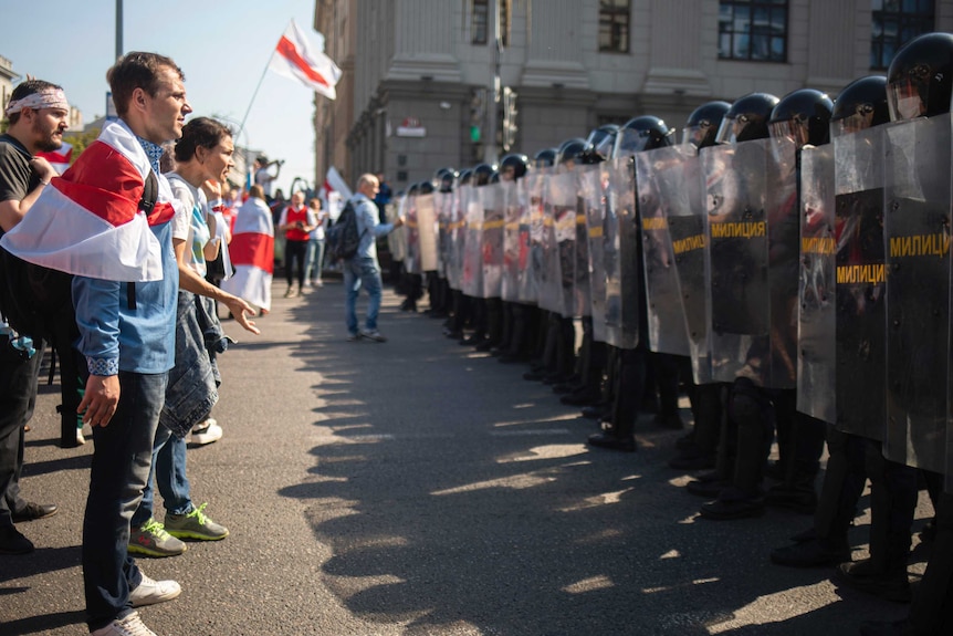 Protesters wearing red and white stand in a line facing a row of armed police holding riot shields.