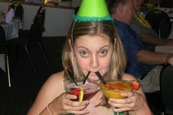 A young woman wearing a party hat, holding two alcoholic drinks with straws.