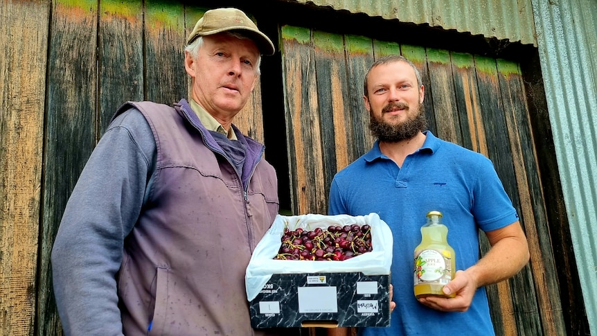 Two man stand beside each other, one holding a box of cherries and the other a bottle of juice.