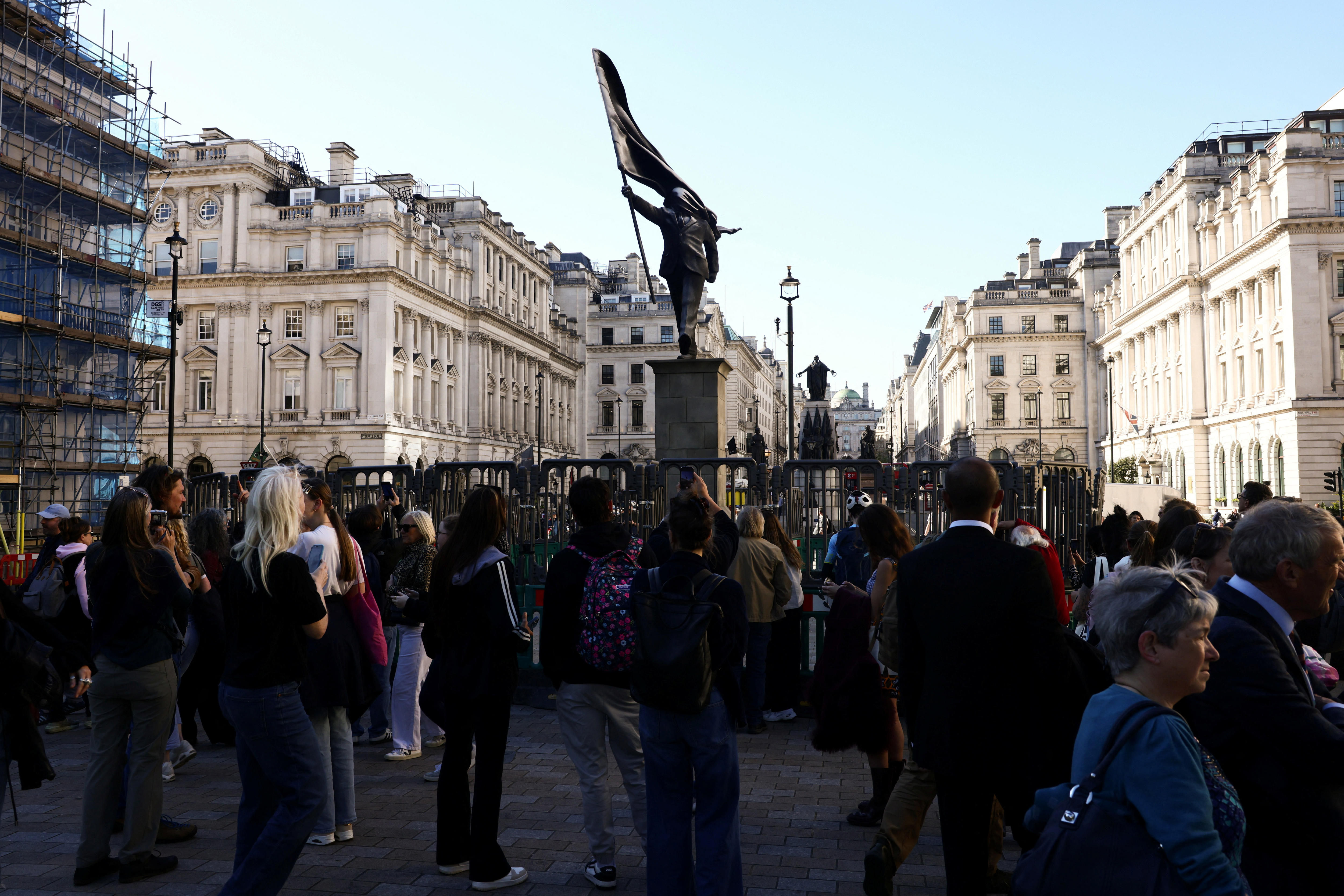 La gente se reúne junto a una estatua que representa a una persona levantándose de un pedestal con una bandera frente a su rostro.