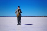 James Valentine stands on a salt flat with a blue sky in a grey suit and plays saxophone.