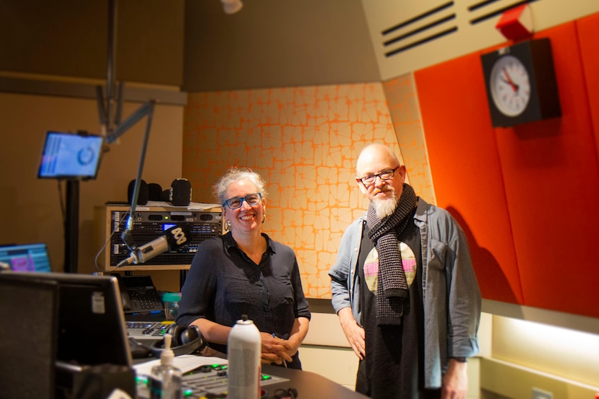 Hilary Harper, with glasses and wide smile, stands next to Michael Mackenzie, with glasses and slight smile, in radio studio.