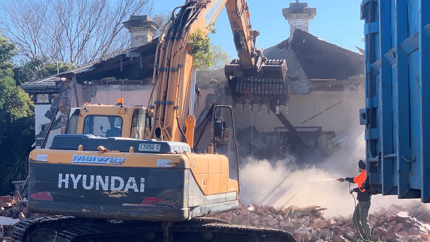 A bulldozer knocking down an old house as a man in high-vis sprays water on the dust.