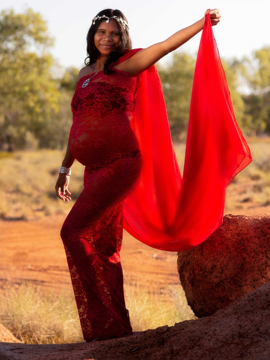 A pregnant Indigenous woman poses on a rock hold out part of her red gown with one hand