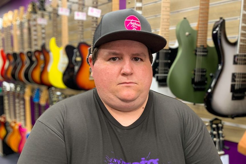 Jake sits in a music store with guitars behind wearing a cap and looking at the camera with a serious expression