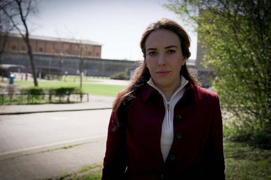 Stella wears a red coat and looks at the camera. The high walls and chain fence of the prison is in the background behind her.