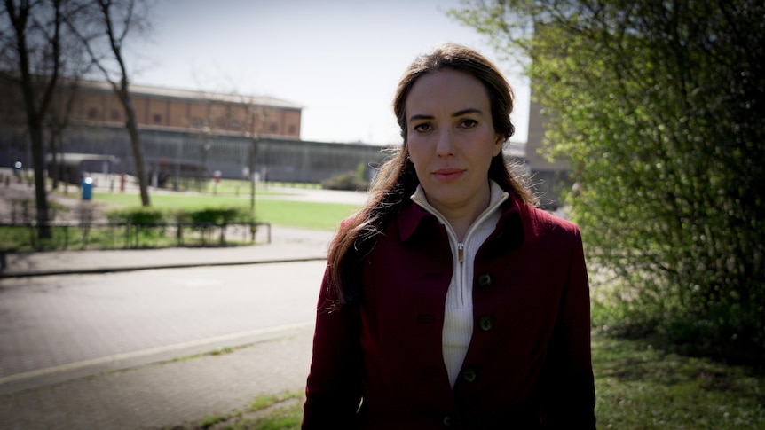 Stella wears a red coat and looks at the camera. The high walls and chain fence of the prison is in the background behind her.