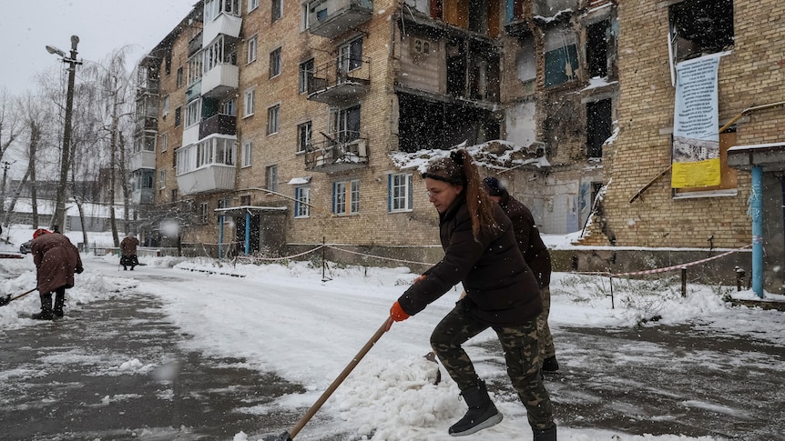 A woman shovels snow in front of a brown building. 