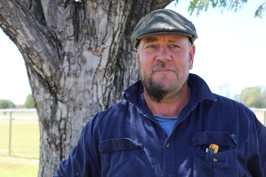 a man stand underneath a tree in a blue uniform with half of his beard shaved off and one eyebrow missing