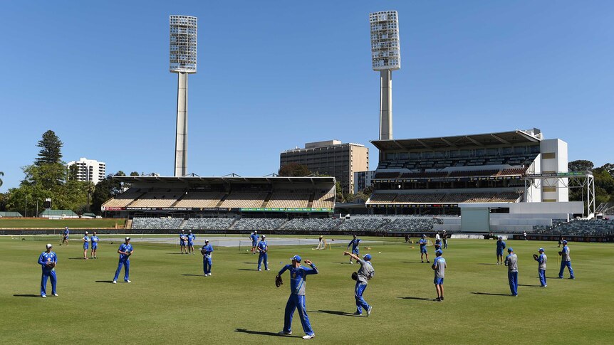 A wide shot of the Australian cricket team training on the WACA ground with two light towers in the background.