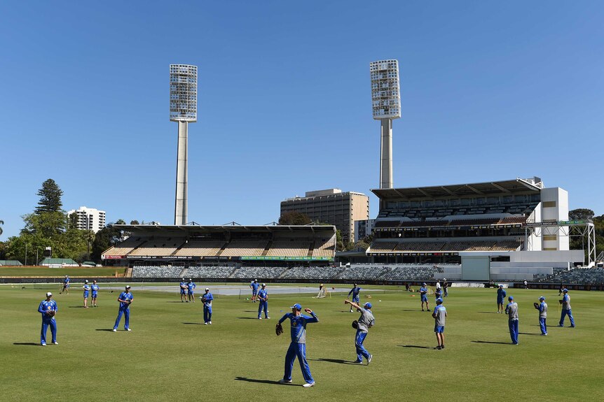 A wide shot of the Australian cricket team training on the WACA ground with two light towers in the background.