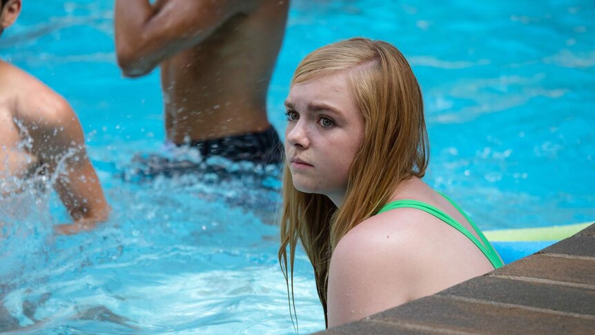 13-year-old in bright green one-piece swim suit sitting looking anxious in swimming pool two male bodies in background.