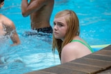 13-year-old in bright green one-piece swim suit sitting looking anxious in swimming pool two male bodies in background.