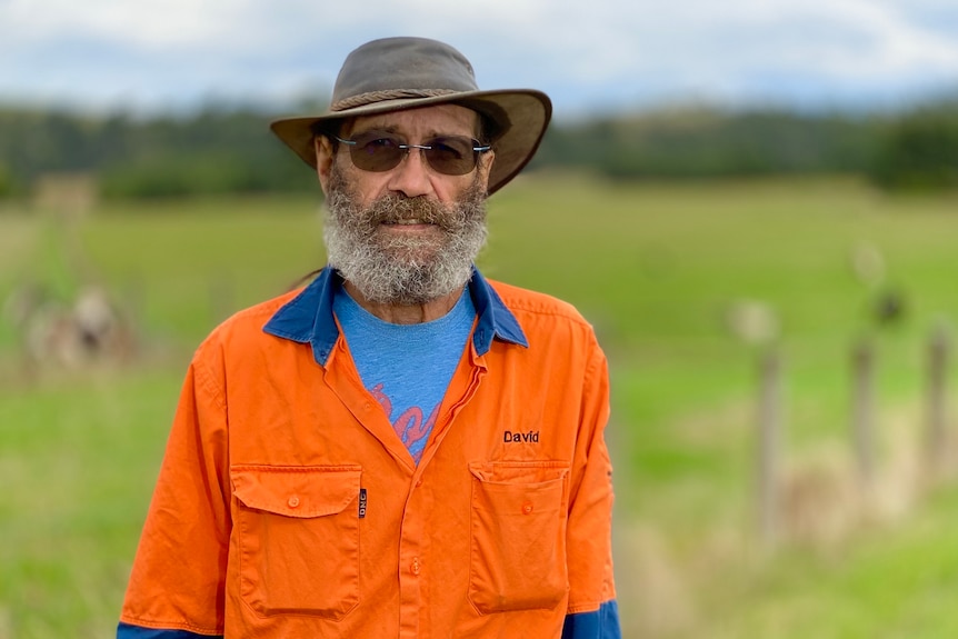 Dave Wandin stands in front of a field on Coranderrk Station, wearing an orange jacket.