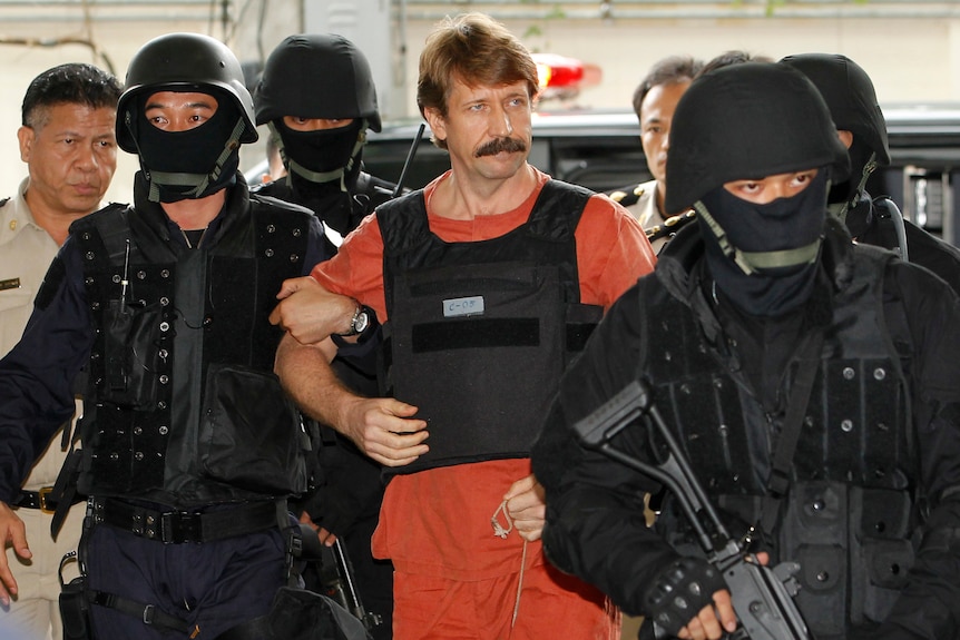A man in an orange prison outfit and flak jacket is escorted by police in black military gear