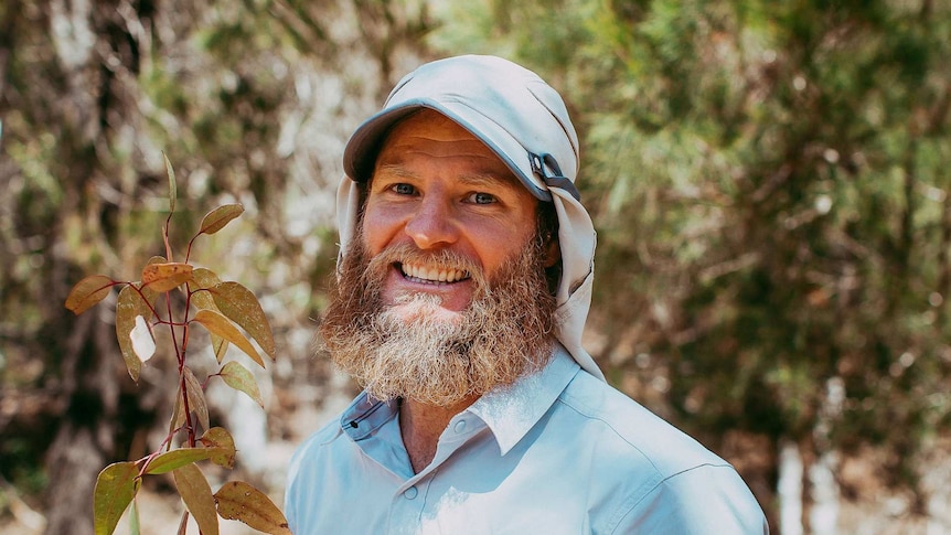 This is a photo of a bearded man in a legionnaires cap standing holding a plant in the bush