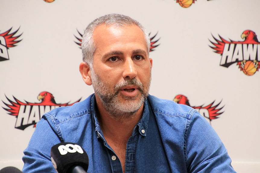 Dorry Kordahi sits and talks in front of a Hawks media wall, wearing a blue button up shirt.