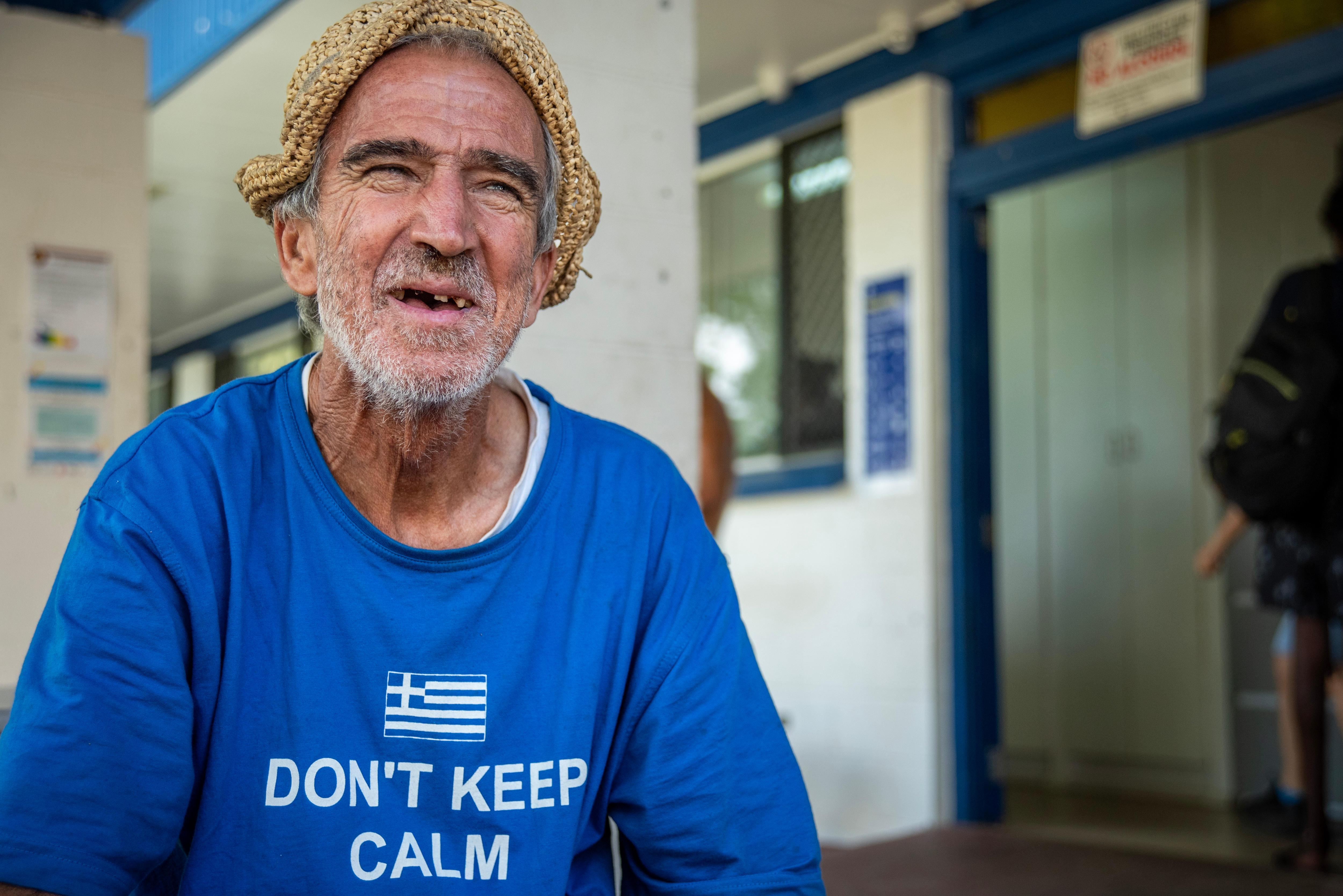 An elderly man in a blue shirt wearing a hat smiles with few teeth. 