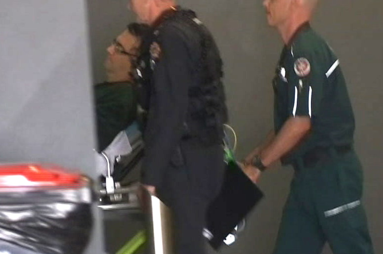 A man is wheeled into hospital on a stretcher with a police guard and paramedic next to him.