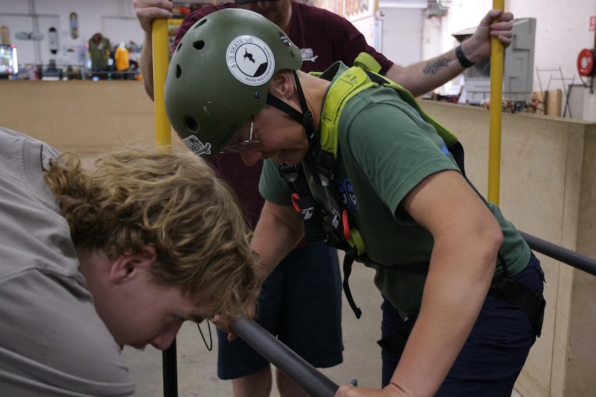 A woman in a green helmet and tshirt laughs as her harness is clipped to her skate frame. 