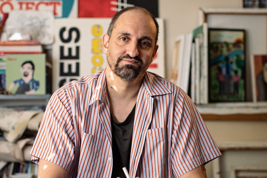 A man with a goatee sits behind a drawing desk, hands clasped around a marker, art lining the walls and shelves behind him