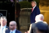 Donald Trump stands at the entrance of Trump tower, a serious expression on his face