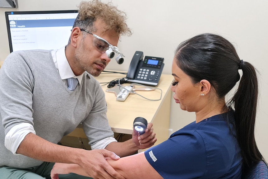 A man wearing magnifying glasses inspects a woman's arm with a light 