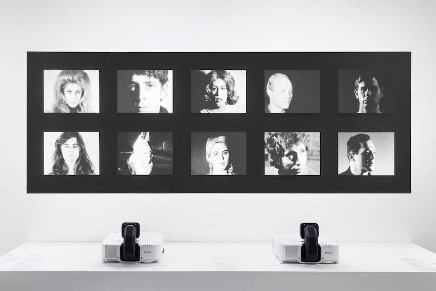 A gallery space, showing two projectors beaming 10 videos onto the wall: close-ups of celebrities including Lou Reed