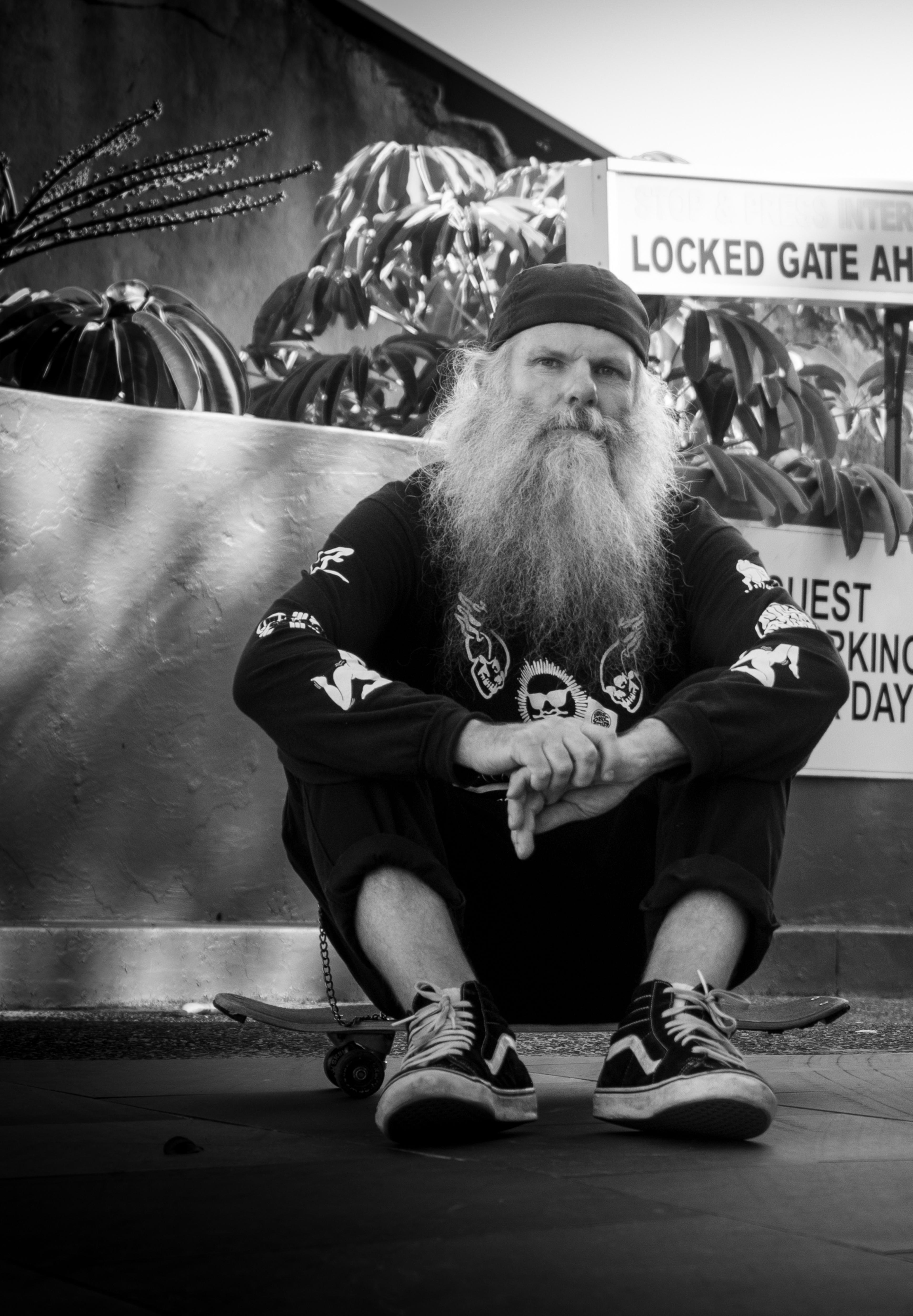 A black and white photo of Shaun Gladwell, a white man with long grey hair and beard, wearing a black cap backwards and sitting.