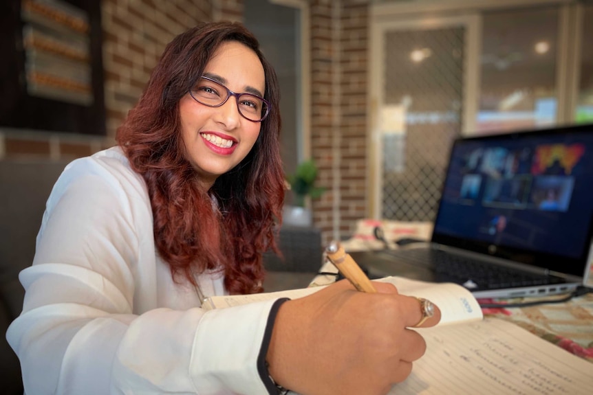 A woman wearing glasses smiles as she sits in front of a computer and writes in a book.