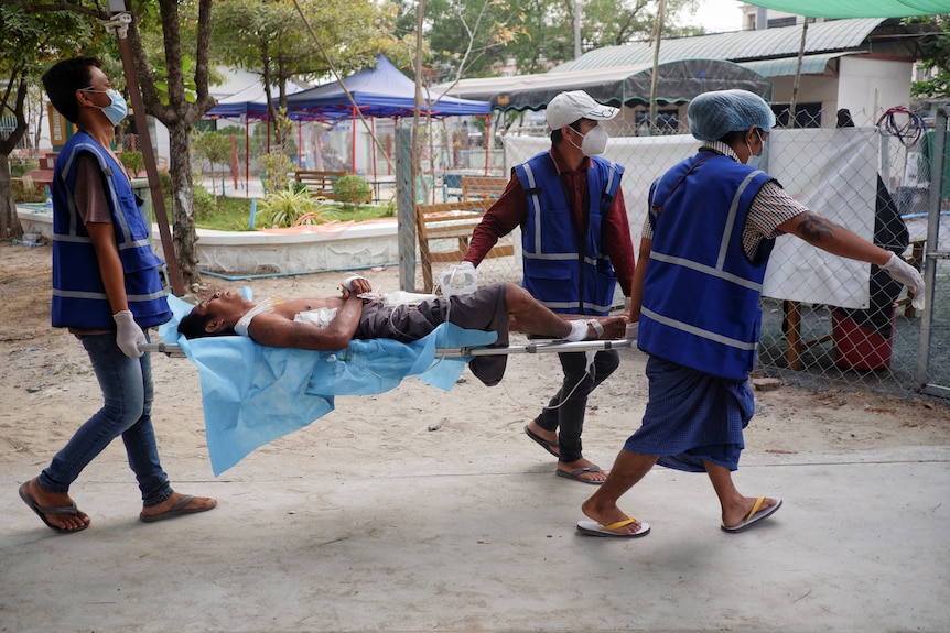 Man lies on stretcher carried by 2 others.
