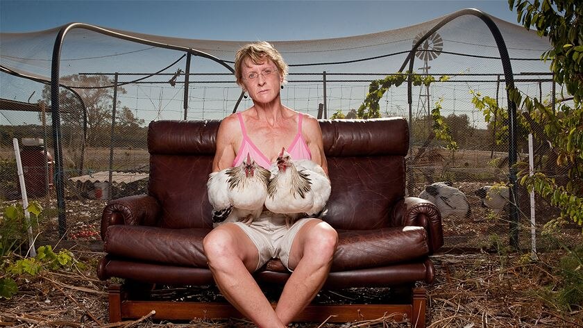 Lorna Thompson sitting on a leather couch in front of a vegie patch, holding two large chickens on her lap
