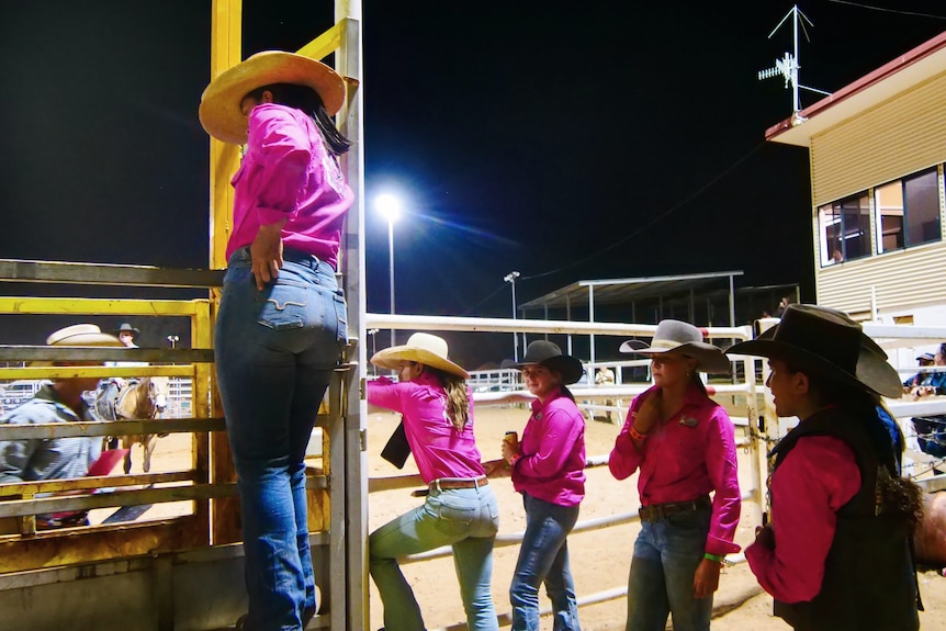 Five women in pink shirts and cowboy hats stand behind a rodeo arena
