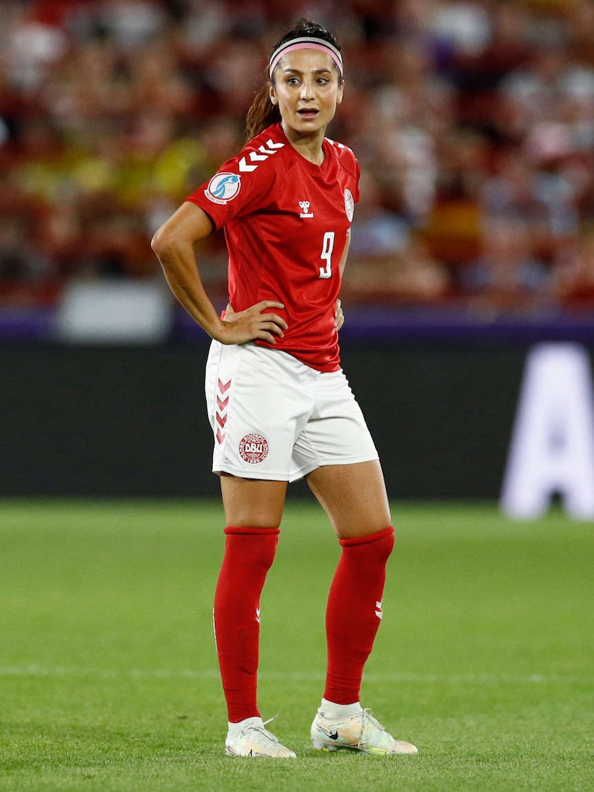 A female soccer player wearing red and white holds her hands on her hips and looks on during a game