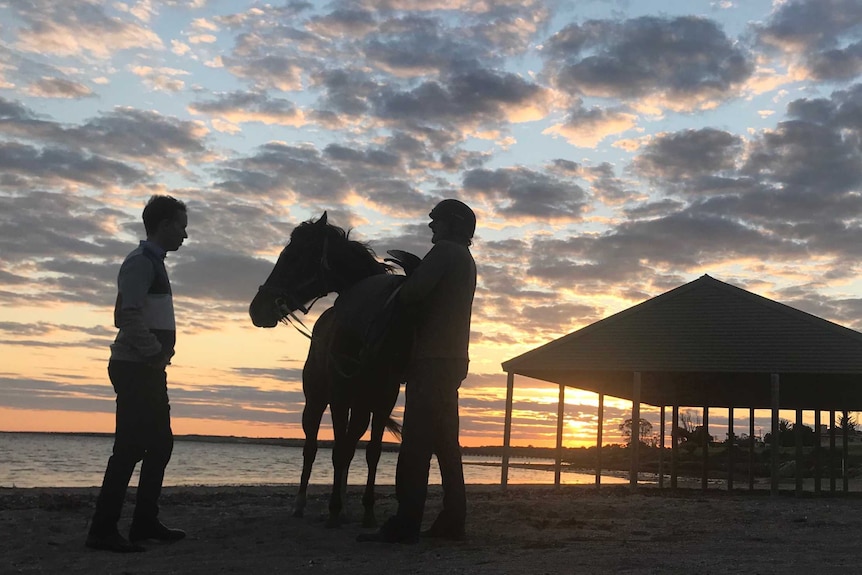 Jockey and trainer stand with horse at sunrise on the beach