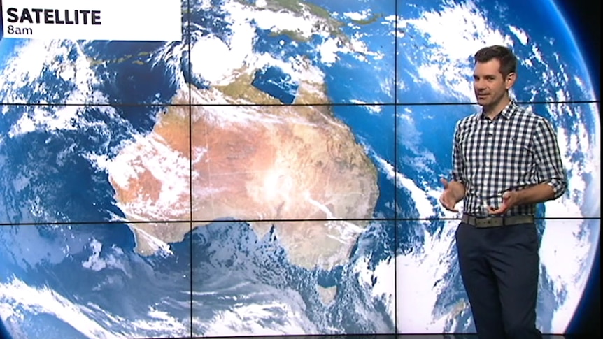 Weather presenter in checked shirt stands in front of synoptic chart map of Australia backdrop on set.
