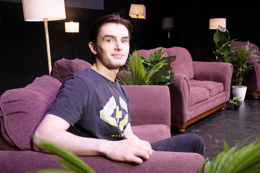 Samuel sits on a chair on a stage wearing a dark purple t-shirt.