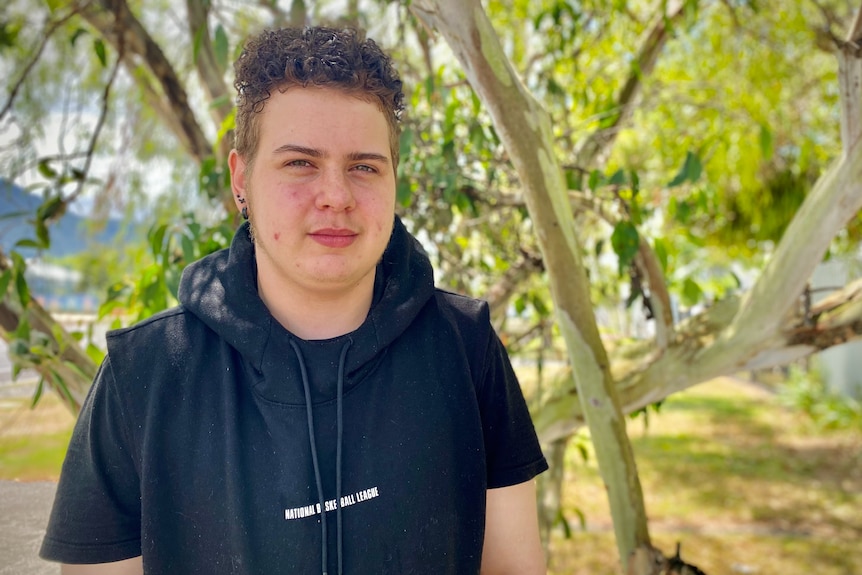 A man in a black sweatshirt stands in front of a tree in a park 