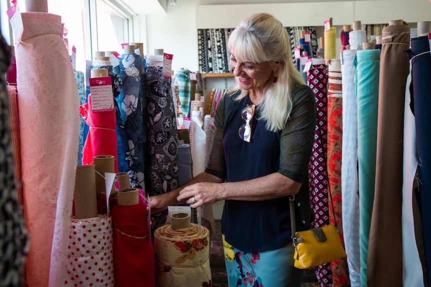 Mary Dobbie stands in a room of fabric rolls, arms reached out touching some red material.