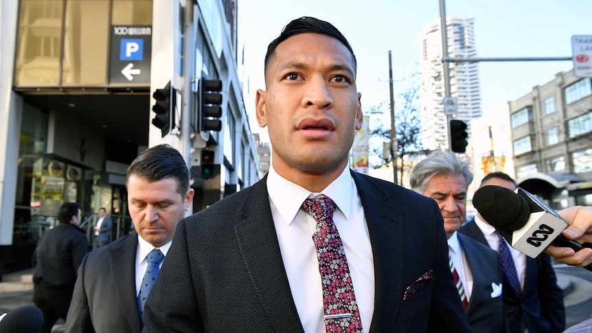 Israel Folau wears a suit and crosses the street while microphones are pointed at him