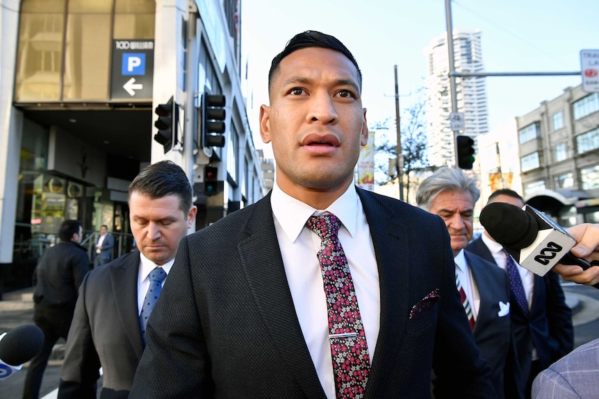 Israel Folau wears a suit and crosses the street while microphones are pointed at him