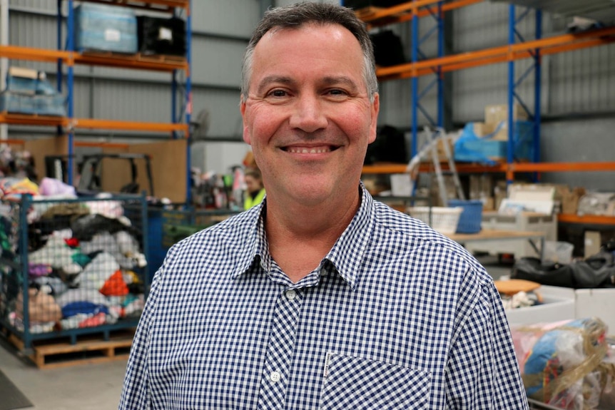 A head and shoulders shot of a smiling middle aged man standing in a warehouse with a checked shirt on.