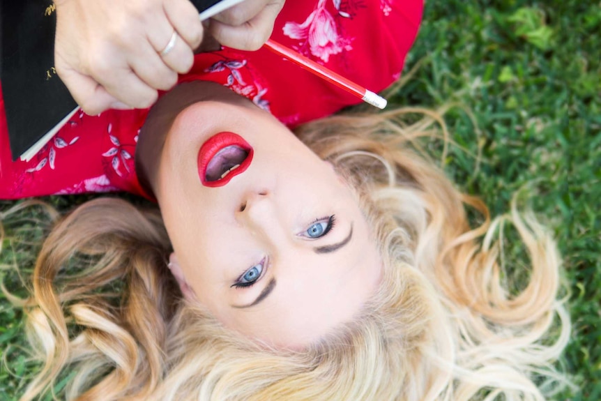 Woman holds a notebook and pencils and stares into camera while lying in grass.