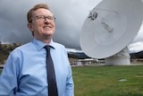 A man in a blue shirt and glasses stand in a field in front of a huge antenna. 
