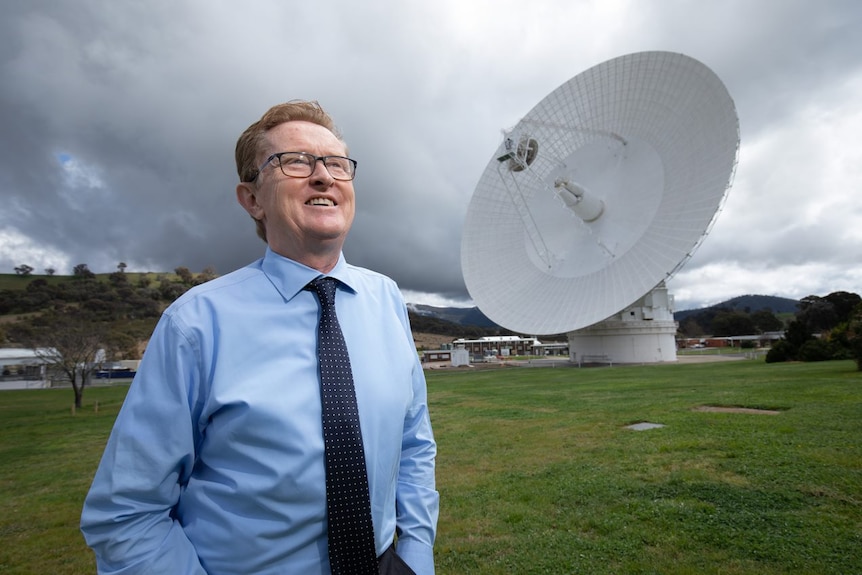 A man in a blue shirt and glasses stand in a field in front of a huge antenna. 