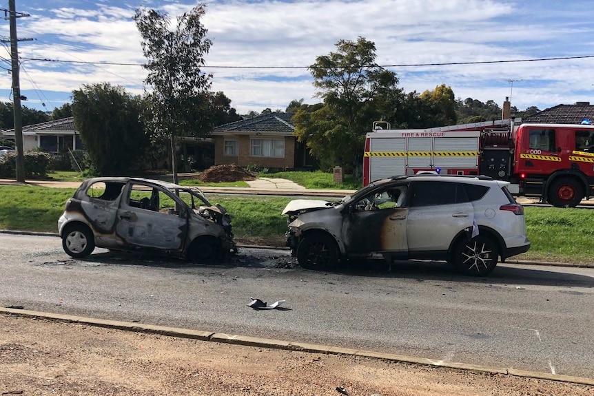 Two burnt out cars facing each other in the middle of a road, with a fire engine in the background.