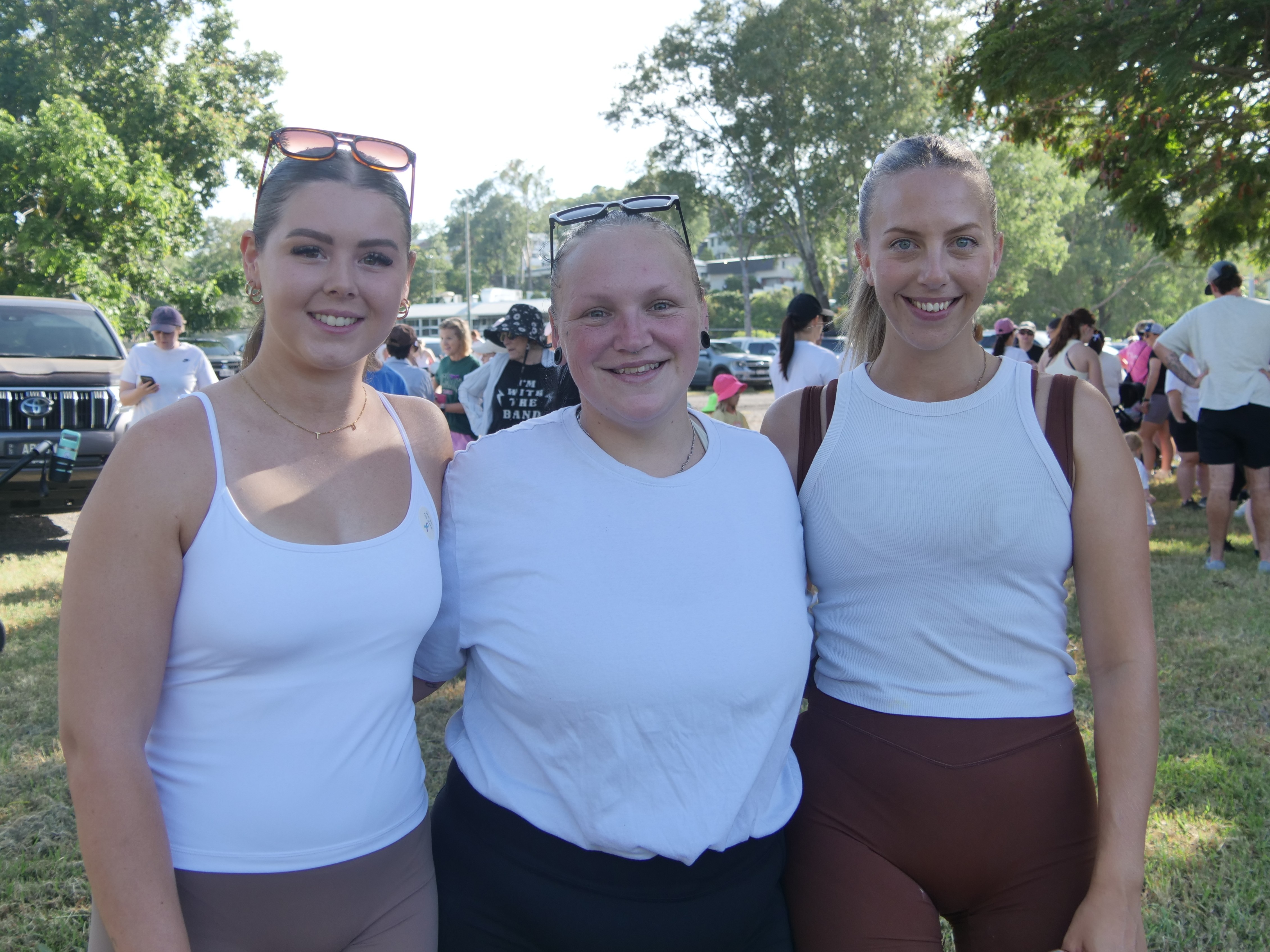 Tres mujeres jóvenes con blusas blancas sonriendo a la cámara, multitud de personas al fondo, árboles, día soleado.