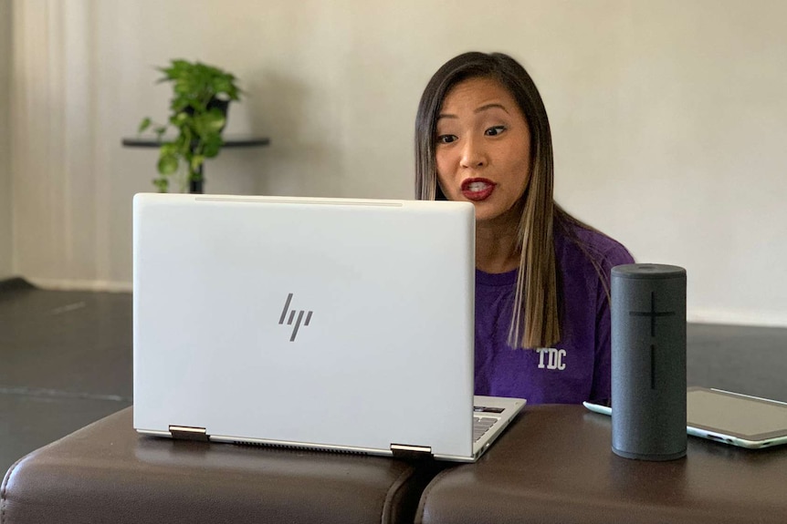 A woman sits at a desk in a room in front of a laptop and bluetooth speaker talking during a virtual dance class.